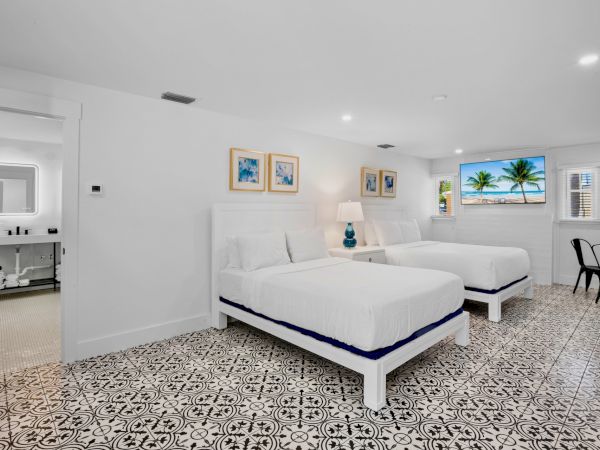 A bright bedroom with two white beds, decorative floor tiles, paintings, a lamp, and a window with a scenic view completes the space.