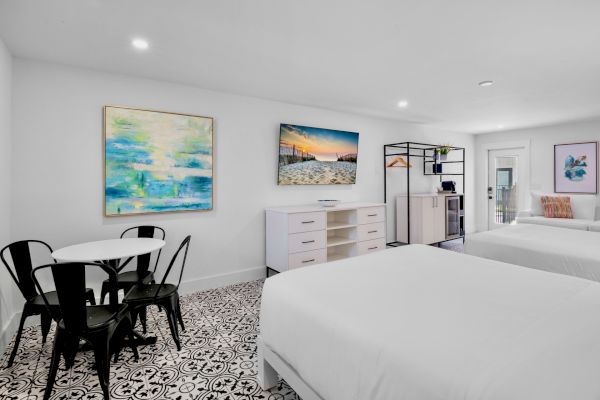 The image shows a modern hotel room with two beds, a table with chairs, art on the walls, and patterned flooring.