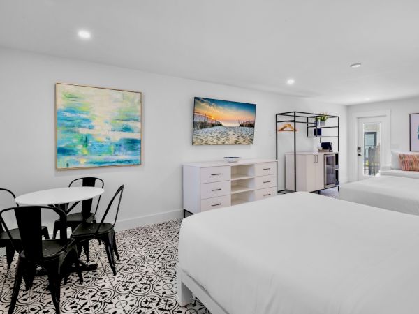 The image shows a modern hotel room with two beds, a table with chairs, art on the walls, and patterned flooring.