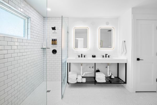 A modern bathroom with white tiles, double sinks, illuminated mirrors, glass shower, and neatly stacked towels underneath.
