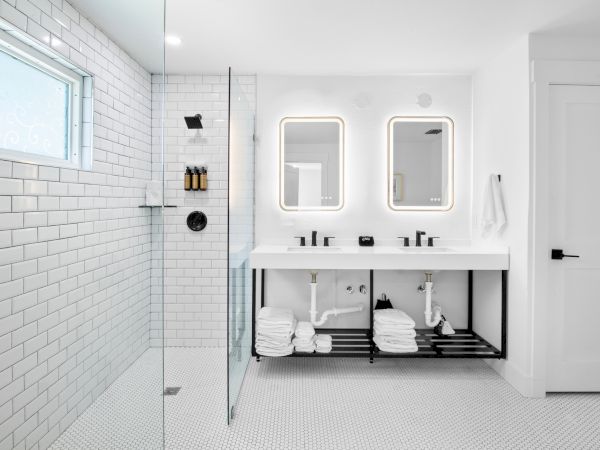 A modern bathroom with white tiles, double sinks, illuminated mirrors, glass shower, and neatly stacked towels underneath.