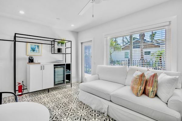 The image shows a bright living room with a white couch, decorative pillows, patterned floor, and a shelving unit with a mini fridge and plants.