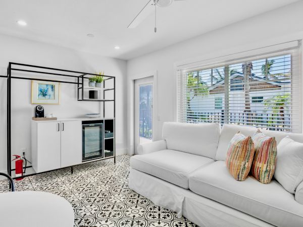 The image shows a bright living room with a white couch, decorative pillows, patterned floor, and a shelving unit with a mini fridge and plants.