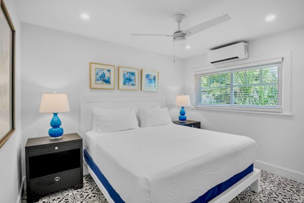 A bright bedroom with a large bed, blue lamps, artwork, and a window. A ceiling fan and air conditioner are also visible.