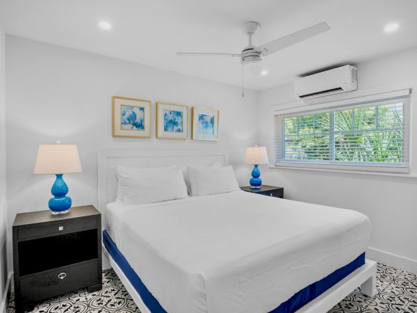 A bright bedroom with a large bed, blue lamps, artwork, and a window. A ceiling fan and air conditioner are also visible.