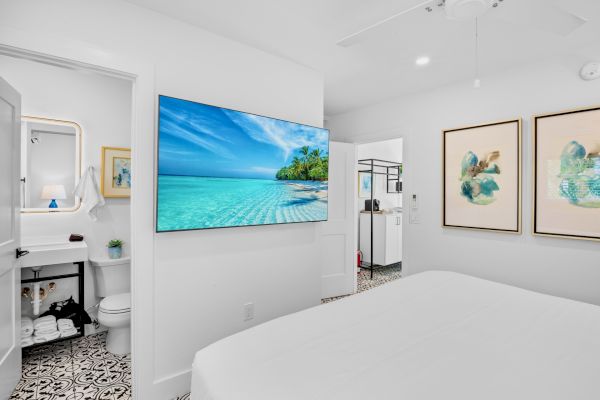 A bright bedroom with a large TV showing a beach scene, adjacent bathroom, patterned floor tiles, and abstract wall art.