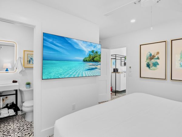 A bright bedroom with a large TV showing a beach scene, adjacent bathroom, patterned floor tiles, and abstract wall art.