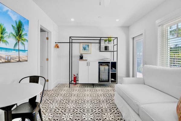 A modern living room with patterned floor tiles, a white couch, a small dining area, a mini fridge, and a beach-themed wall art.