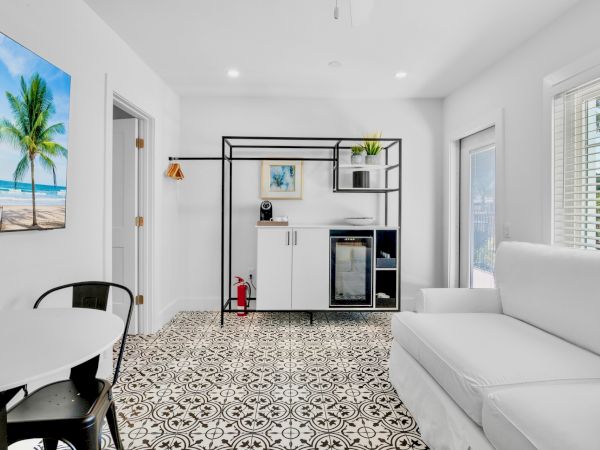 A modern living room with patterned floor tiles, a white couch, a small dining area, a mini fridge, and a beach-themed wall art.