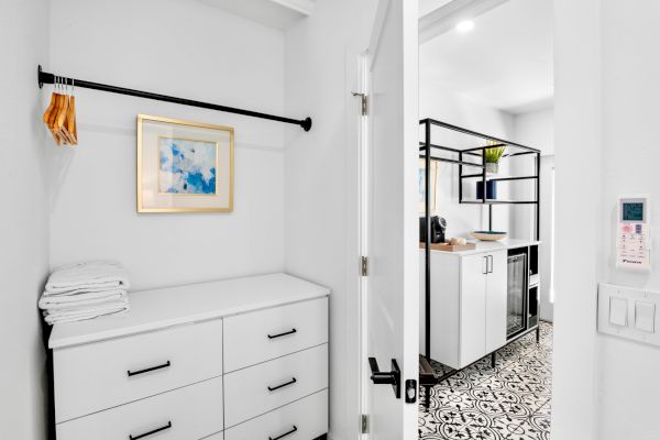 The image shows a white closet with drawers, towels, hangers, and an open door leading to a room with patterned flooring.