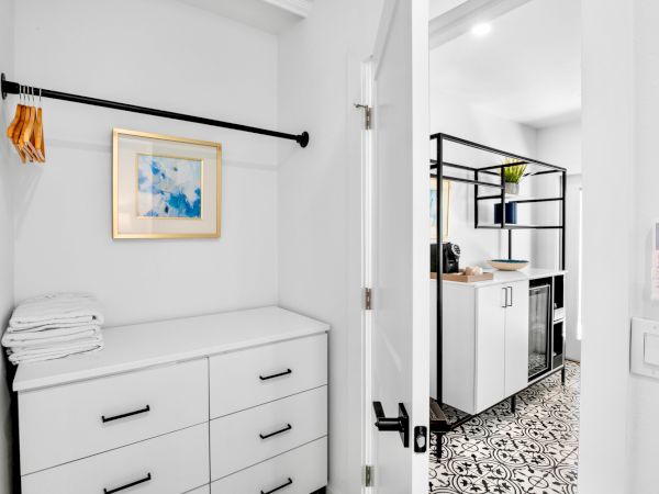 The image shows a white closet with drawers, towels, hangers, and an open door leading to a room with patterned flooring.