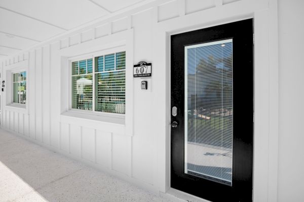 The image shows a white exterior wall with a black-framed door and windows, all featuring closed blinds, with a room number sign displayed.