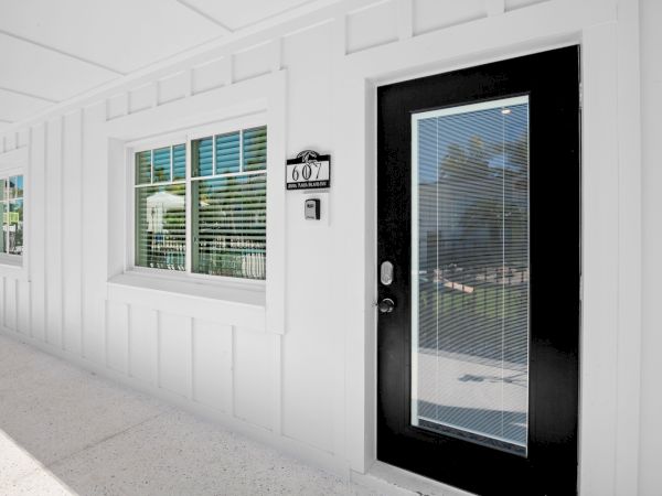The image shows a white exterior wall with a black-framed door and windows, all featuring closed blinds, with a room number sign displayed.