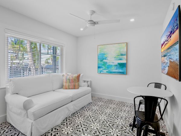 A bright room with a white couch, patterned floor, wall art, round table with chairs, ceiling fan, and a TV displaying a beach scene.