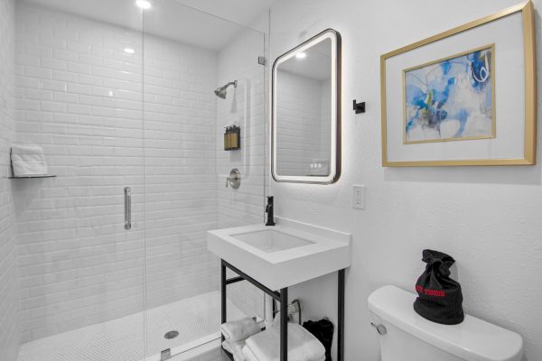 A modern bathroom with glass shower, illuminated mirror, sink, and artwork above the toilet. Shelving and visible towels are included.