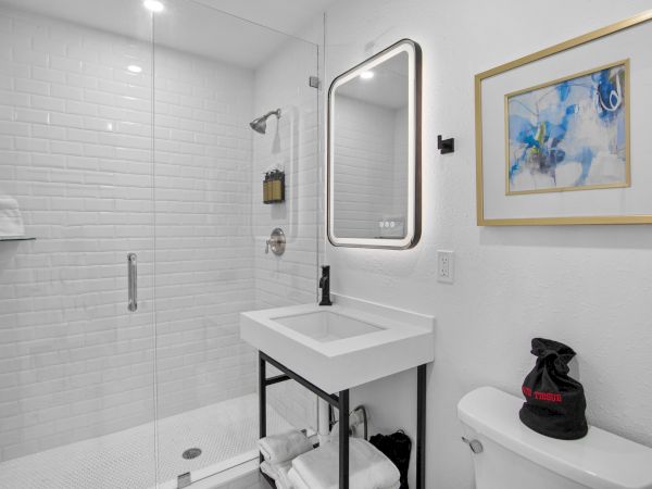 A modern bathroom with glass shower, illuminated mirror, sink, and artwork above the toilet. Shelving and visible towels are included.