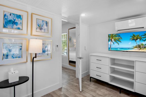 A modern room with tropical wall art, a beach view on TV, white drawers, a floor lamp, and a black side table with a tissue box.