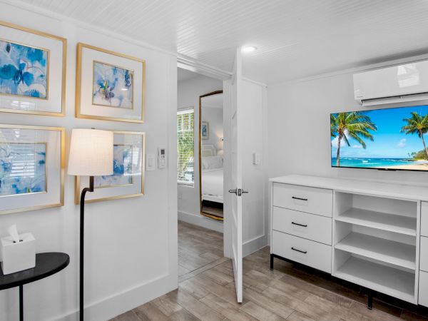 A modern room with tropical wall art, a beach view on TV, white drawers, a floor lamp, and a black side table with a tissue box.