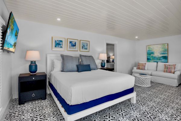 A modern bedroom featuring a bed, artwork, a sofa, patterned floor, TV, lamps, and a table, all in a bright and stylish decor.