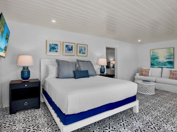 A modern bedroom featuring a bed, artwork, a sofa, patterned floor, TV, lamps, and a table, all in a bright and stylish decor.