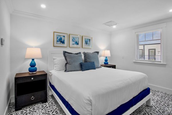 A neatly arranged bedroom with a bed, blue-themed lamps, artwork on the wall, and patterned flooring.