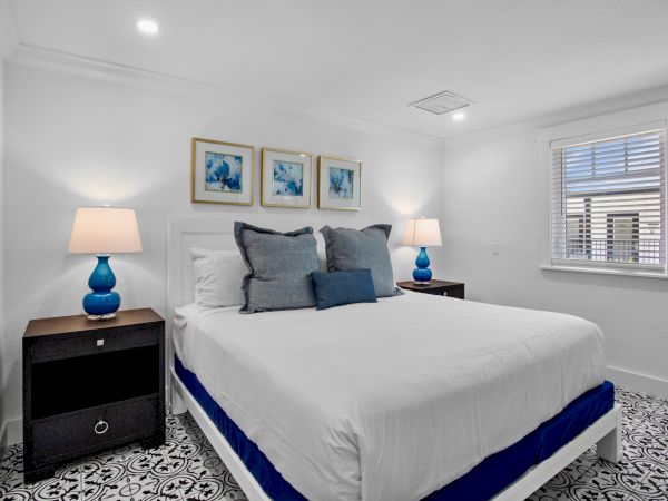 A neatly arranged bedroom with a bed, blue-themed lamps, artwork on the wall, and patterned flooring.