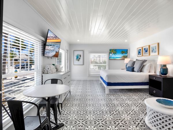 A bright, modern bedroom with a patterned floor, a bed, a TV, and a small dining area by the window. Coastal decor enhances the setting.