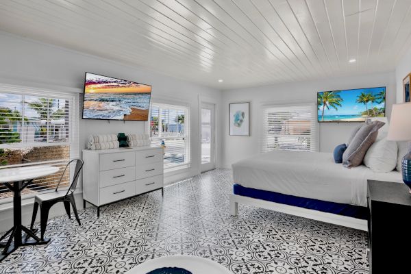 A bright bedroom with patterned flooring, a bed, two TVs, a dresser, and a small table with a chair by the windows.