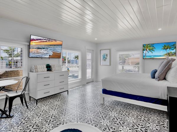 A bright bedroom with patterned flooring, a bed, two TVs, a dresser, and a small table with a chair by the windows.