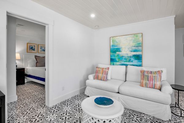 A small living room with a white sofa, colorful pillows, abstract art, and patterned floor tiles. Adjacent is a partially visible bedroom.