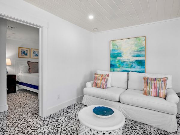A small living room with a white sofa, colorful pillows, abstract art, and patterned floor tiles. Adjacent is a partially visible bedroom.