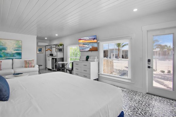 A bright, modern bedroom with a bed, sofa, TV, decorative cushions, artwork, large window, and a door leading outside.