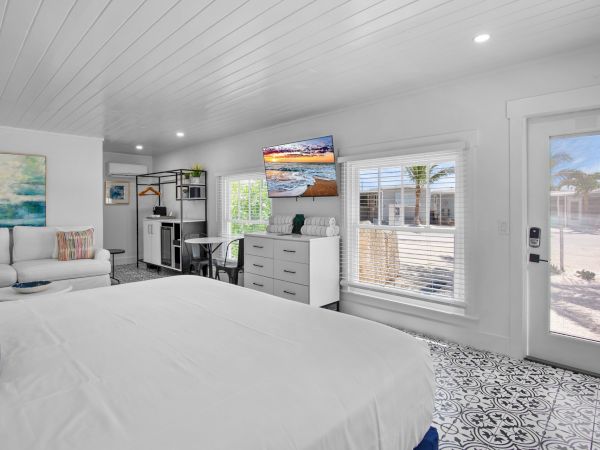 A bright, modern bedroom with a bed, sofa, TV, decorative cushions, artwork, large window, and a door leading outside.