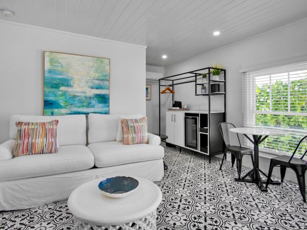 A modern living room with a white sofa, colorful pillows, abstract painting, round table, and chairs, featuring black and white patterned floor.