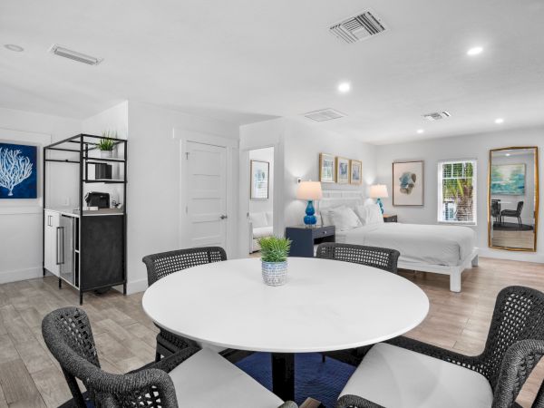 Modern open-plan bedroom with kitchenette, dining table, and artwork; bright and airy with neutral tones.
