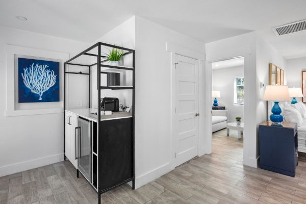 A modern, minimalist room with light wood floors, a sleek kitchenette, blue artwork, and a view into a bright, white adjoining room.