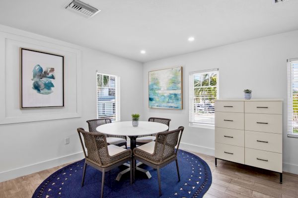A modern dining room with a round table, four chairs, blue rug, abstract wall art, and a white chest of drawers by the windows.