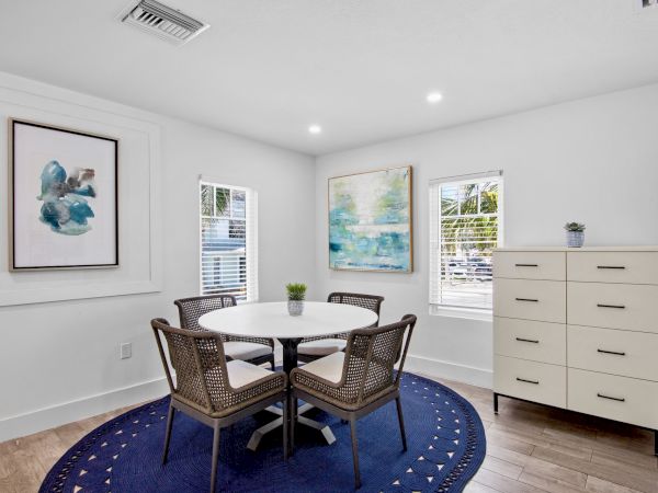 A modern dining room with a round table, four chairs, blue rug, abstract wall art, and a white chest of drawers by the windows.