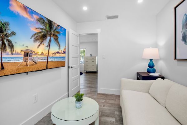 A modern living room with a white couch, glass table, beach scene on TV, lamp on side table, and open door to another room’s glimpse.
