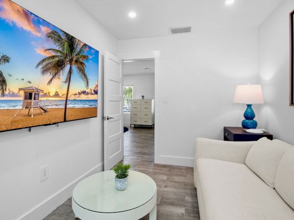 A modern living room with a white couch, glass table, beach scene on TV, lamp on side table, and open door to another room’s glimpse.