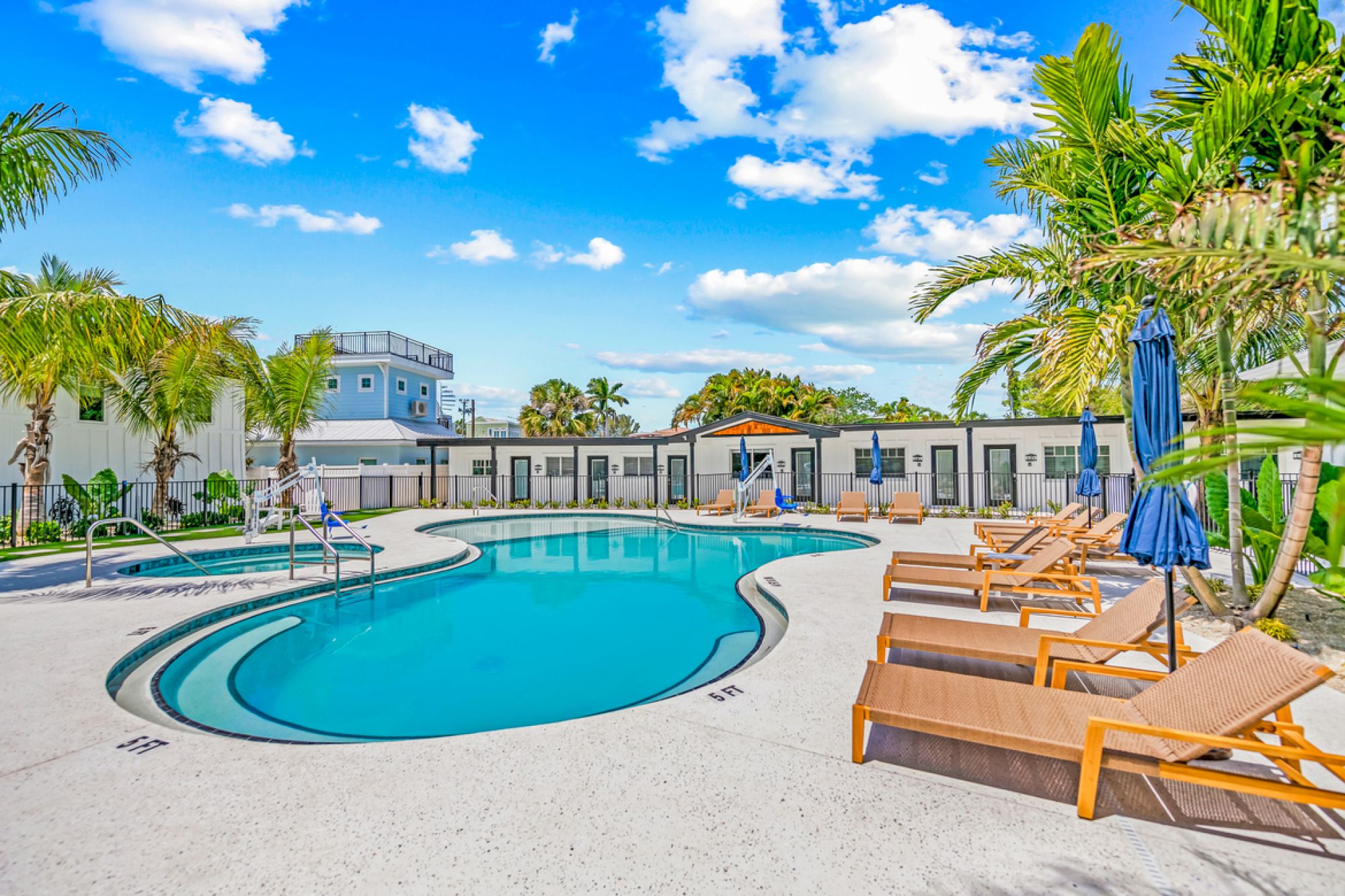 The image shows a serene outdoor swimming pool area with lounge chairs, umbrellas, palm trees, and a bright blue sky with clouds.