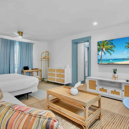 Bright bedroom with a bed, blue curtains, dresser, rattan coffee table, ceiling fan, and a framed tropical beach scene.