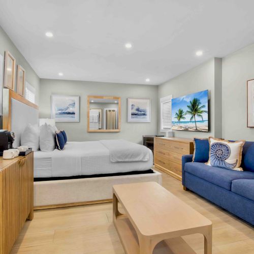 Bright modern living room with a white sofa-bed, blue couch, coffee table, framed beach art, windows, and wall air conditioner.