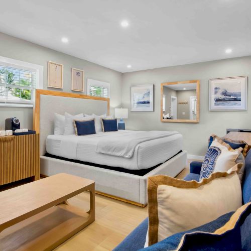 A bright modern bedroom with a neatly made bed, sofa and coffee table, framed art, and large windows.