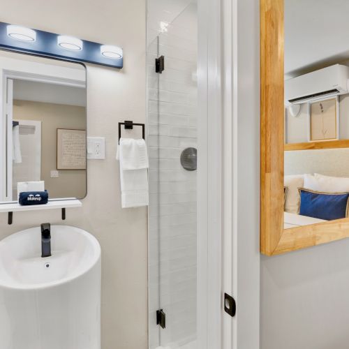 The image shows a bathroom with a sink, mirror, and shower, next to a small bedroom with a bed and a wooden-framed window between them.