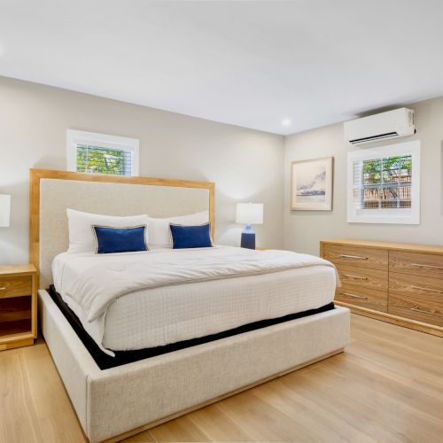 A modern bedroom with a double bed, two bedside tables, lamps, artwork, a dresser, and an air conditioner on a light wooden floor.