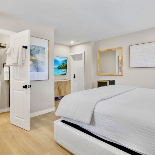 A neatly arranged bedroom with a bed, white linens, bedside tables, artwork, and an open door leading to a bathroom with a sink and towels.