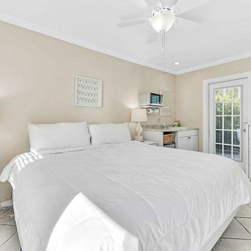A cozy bedroom with a large bed, two white nightstands, a dresser, and a tiny kitchenette near a door leading outside.