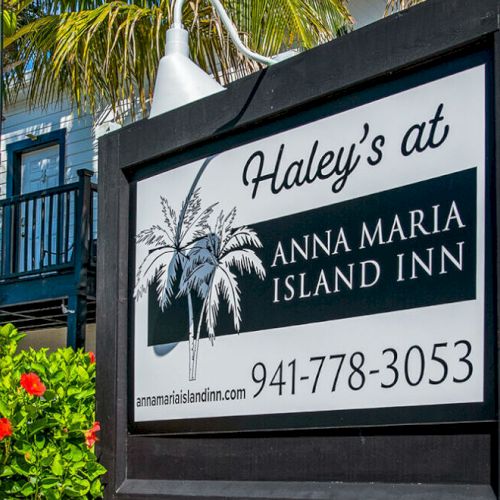 A sign for “Haley’s at Anna Maria Island Inn” with contact info, surrounded by tropical plants and palm trees.
