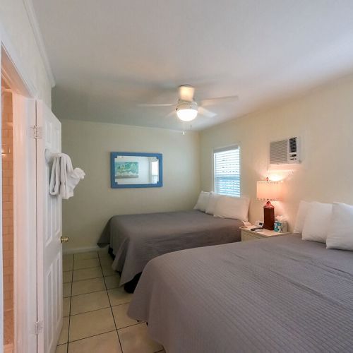 A cozy bedroom with two beds, ceiling fan, lamp on nightstand, art on wall, and door to bathroom with towel rack. Bright and clean ambiance.